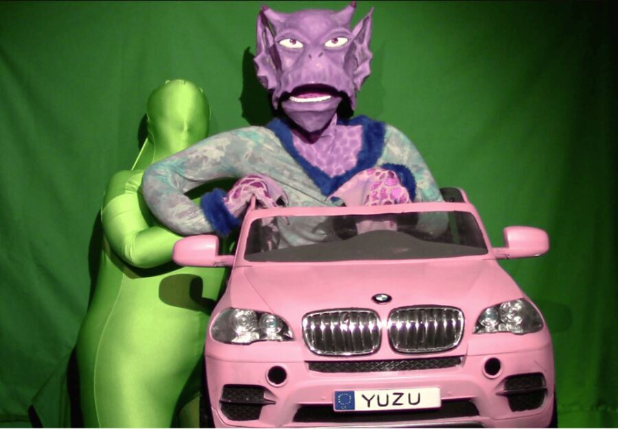 A lilac dragon-headed puppet sitting in a pink toy car on a green screen set.
