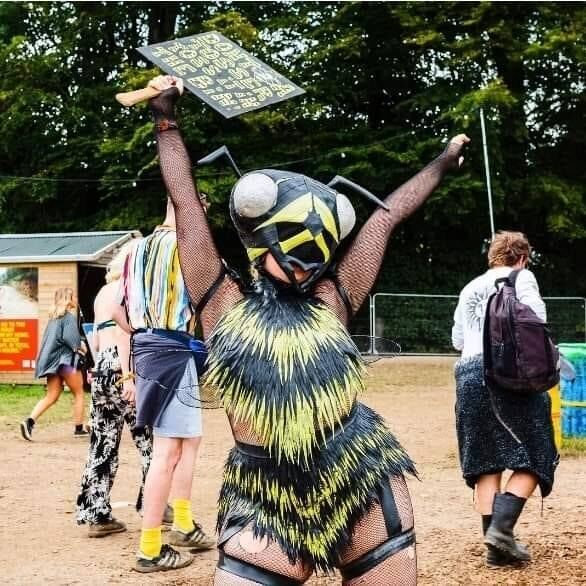 A performer in a black and yellow bee costume with wings and antennae, holding a placard aloft at a festival.