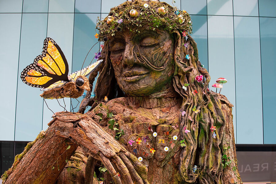A towering figure built from bark, moss and flowers, a butterfly resting on its outstretched hand.