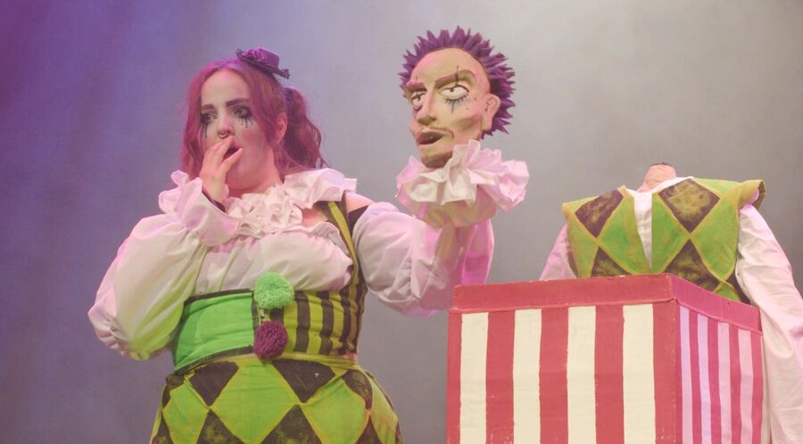 Two harlequin figures in green and pink costume beside a striped box, one a performer and one a puppet.