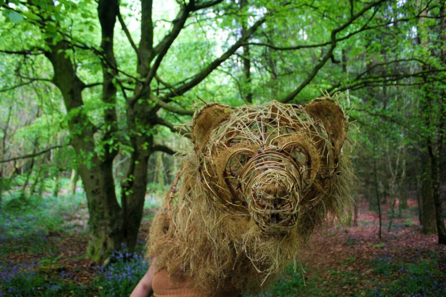 A bear's head woven from willow, standing among trees and bluebells in woodland.